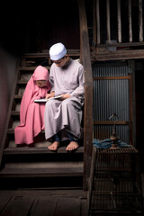 A brother and a cute little girl, an Asian Muslim, is practicing reading Quran with faith on the stairs in an old wooden house.
