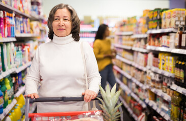 Pensive middle aged female shopping at store, walking among shelves and choosing products