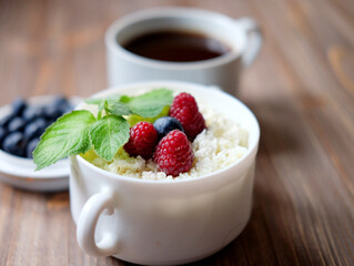A cup of coffee and Cottage cheese in white bowl with raspberries and blueberries on wooden background.