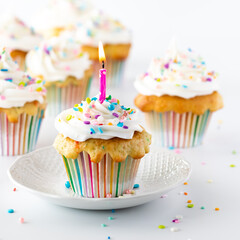 Close up of a confetti cupcake with a pink lit candle with other cupcakes in soft focus in behind. Instagram crop.