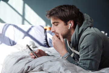 Man looks at his phone wearing headphones in bed