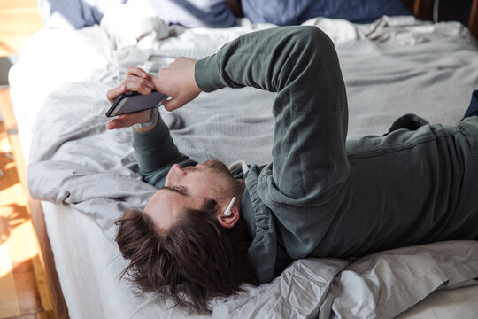 Man looks at his phone wearing headphones in bed