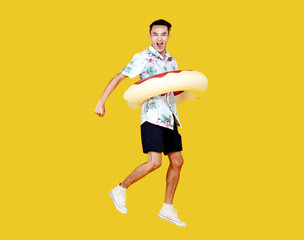 Fototapeta na wymiar Young attractive Asian man wearing hat and white Hawaiian shirt and donut swim ring around his waist jumping with excitement against yellow background. Concept for beach vacation holiday
