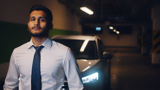 Indian Man Standing In The Parking Lot With His Car Parked Behind Him. Man Dressed In Formal White Shirt And Blue Tie With Serious Facial Expressions. Concept Of Work Load. Night Time