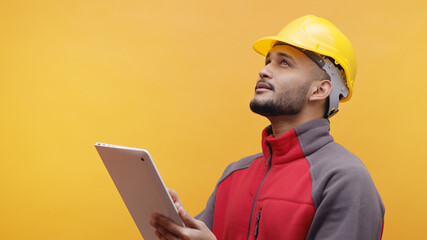 A young Engineer wearing yellow safety helmet holding a tablet in his hand. Studio shot with yellow background. Engineering and construction concept. 