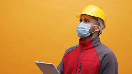 A young Engineer wearing yellow safety helmet and a mask holding a tablet in his hand. Studio shot...