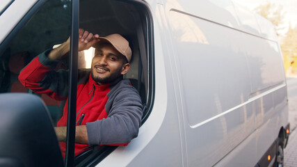 Delivery guy wearing red uniform sitting in the white van tipping his cap. Happy Indian guy sitting in the front seat and smiling at the camera. Courier concept.