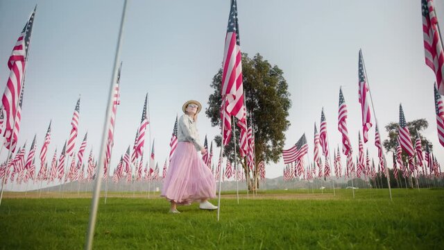 Young woman walking between flagpoles on summer day. Model celebrating Independence or Labor day in the green park with many American flag hanging down. Independence and Freedom concept 4K footage