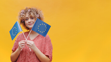 Attractive young girl holding two small European Union Flags in her hands. Studio shot isolated on yellow background. Symbol of Europe, EU association. 
