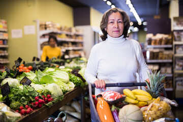 Obraz premium Pensive middle aged female shopping at store, walking among shelves and choosing products