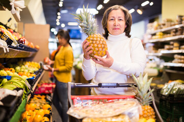 Obraz premium Satisfied elderly woman picking ripe pineapple at grocery supermarket