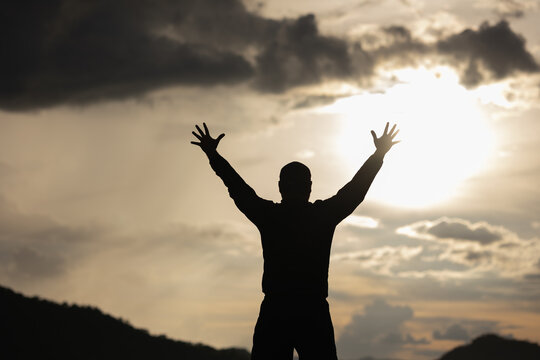 Silhouette Young man raised hand in the air with success goal on sunset background. Man standing on top of the mountain freedom life concept.