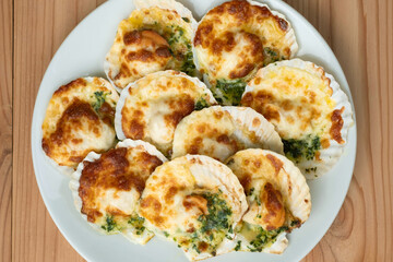 Baked parmesan scallops in a white ceramic plate on a wooden table . Oven baked scallop with cheese, spinach, butter and garlic.