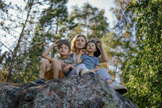 young beautiful woman enjoying spending time with two kids. Elder son kissing mom on cheek. Family is sitting on big rock in Mon Repos Park, Vyborg. Image with selective focus