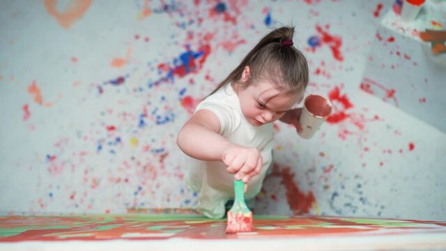 Kid girl with down syndrome draws with a brush on a large canvas in a white room, kid girl with special needs draws a color red-blue abstraction, 4k slow motion.