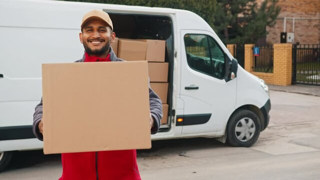 Delivery boy holding a cardboard box in his hands. Smiling and moving the box towards the camera. A white van with stock of cardboard boxes in the background. Concept of parcel delivery service. .