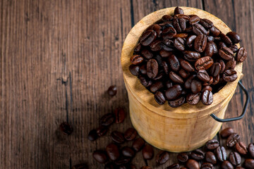 A small wooden bucket of coffee and many coffee beans placed around on a wooden table in a warm, light atmosphere, on dark background, with copy space.
