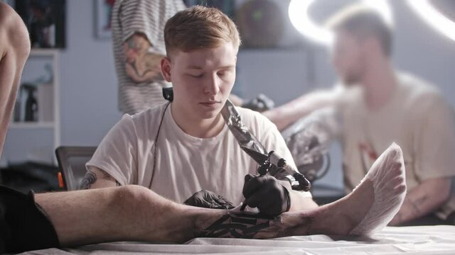 A session at the tattoo salon - young man master tattooing big letters on the leg - looking in the camera