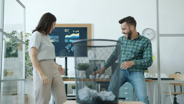 Joyful man and woman playing basketball in office throwing paper balls in basket doing high-five laughing having fun during work break. Job and relaxation concept.