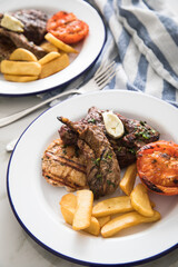 Grillteller mit Rindersteak, Hühnchen Brustfilet, Lamm Filet dazu Steakhaus Pommes, Grilltomate und Kräuterbutter auf Emaille Teller blau weiß, Tuch und Marmor Hintergrund hell 