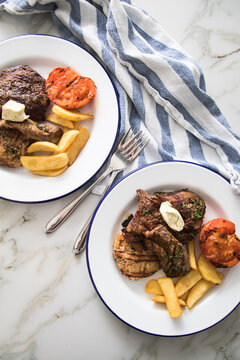 Grillteller mit Rindersteak, H&uuml;hnchen Brustfilet, Lamm Filet dazu Steakhaus Pommes, Grilltomate und Kr&auml;uterbutter auf Emaille Teller blau wei&szlig;, Tuch und Marmor Hintergrund hell 