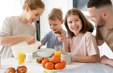 Obraz na płótnie Canvas Young happy family with two cute little kids having breakfast together in kitchen and smiling