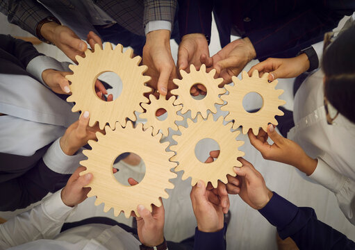 Team of business people connect cogwheels as metaphor for good teamwork, cooperation, partnership and work efficiency. High angle, from above, top view closeup shot of hands holding wooden cogs