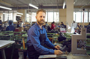 Smiling male shoemaker makes a new pair of leather shoes at a modern shoe factory. Caucasian man who uses a hammer to make holes in a leather material. Concept of working with leather products.