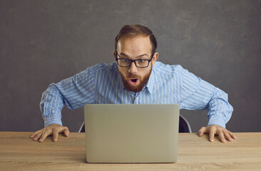 Funny open mouthed nerd in eyeglasses looking at laptop computer screen with big surprise. Shocked...