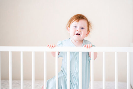 A Toddler With Red Hair Is Holding Onto The Side Of The Crib. Danger For The Child To Fall Out Of Bed. The Baby Learns To Stand At The Support.