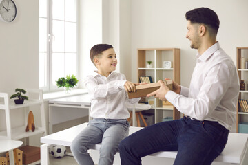 Positive holiday moment. Happy father receives a holiday gift from his loving little son. Family in jeans and white shirts sitting at a table in the room. Gifts for father's day, birthday or men's day