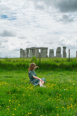 A blonde girl in a green dress sits on a grass of a prairie looking at the ancient monument of Stonehenge at the end of the path