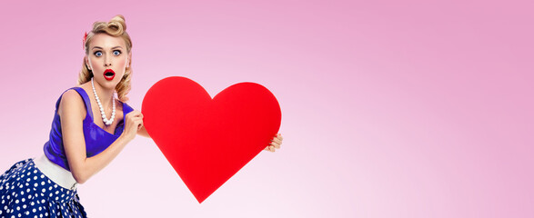 Image of woman holding heart symbol, dressed in pin up style dress with polka dot, over pink background. Caucasian blond model posing in retro fashion and vintage studio concept. Wide.