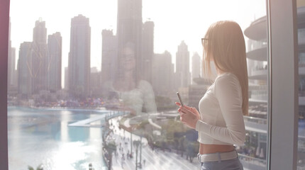 Young woman taking photo at the big window and looking on the Dubai city buildings. Sunset.
