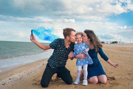 Parents are kissing a little daughter at her cheeks, sitting on the sand near the water. A man holds smoke pellet with blue smoke.