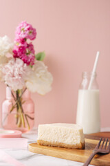 Fototapeta na wymiar Slice of tasty homemade cheesecake with flowers and miilk on pink background. Healthy organic summer dessert pie.