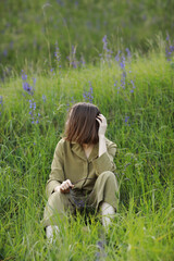 Fototapeta premium Portrait of a young woman sitting in a field on the spring grass among purple flowers. stylish girl enjoys Sunny spring weather. Natural beauty of a woman, natural cosmetics