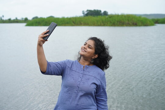 Young female happily taking selfies while Standing Near Lake Medium shot of a pretty Indian girl posing while clicking pictures using a smartphone