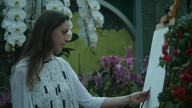 Girl artist in a white dress at work in the flower orchid garden. Doing a pencil sketch, concentrated, focused, hard-working and professional.