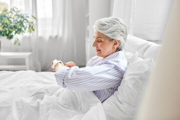 old age, technology and people concept - happy smiling senior woman in pajamas with smart watch sitting in bed at home bedroom