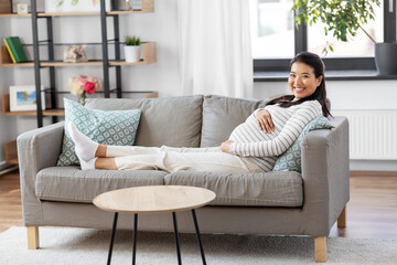 pregnancy, rest, people and expectation concept - happy smiling pregnant asian woman sitting on sofa at home