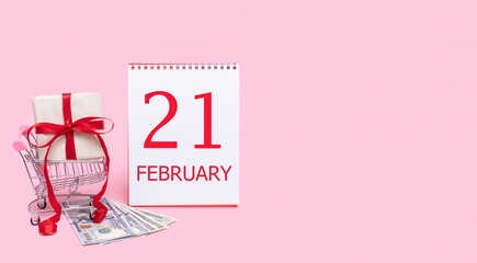 A gift box in a shopping trolley, dollars and a calendar with the date of 21 february on a pink background.