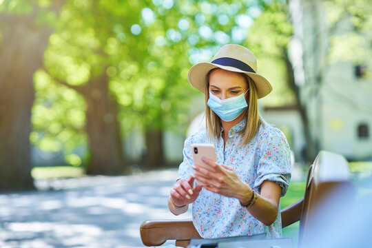 Woman in mask with smartphone sitting on a bench in the park.