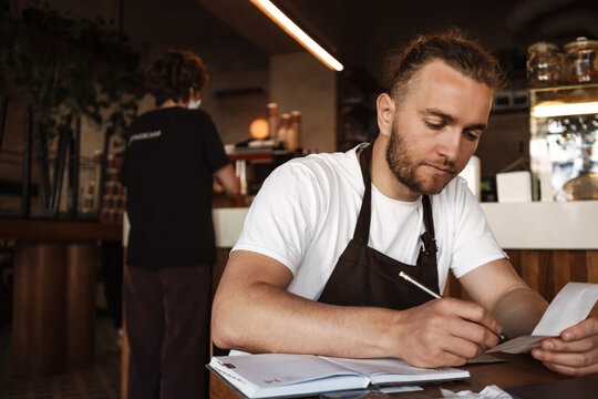 Young white cafe worker in apron writing down notes while sitting at table