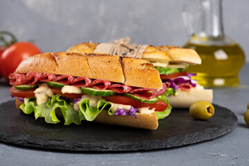 Home-made sandwich of crispy baguette slices with salami, vegetables and lettuce leaves and red cabbage seasoned with a spicy sauce on a gray background.