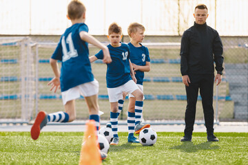 Fototapeta premium Coaching Kids Soccer. Young Man as Soccer Coach Trainer on Football Children Camp. Soccer Boys in Blue Uniforms Running Fast and Kicking Balls on Training Pitch