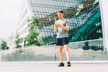 Tired Young man runner standing and using mobile phone and relaxing after sport training. Holding water bottle while doing fitness workout in city urban street, cloudy sky at summer