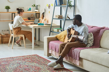 African man playing guitar on sofa with woman sitting at the table and working on laptop in the room at home
