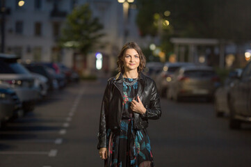 Fototapeta premium a beautiful young woman in a leather jacket walks through a parking lot. Night city portrait