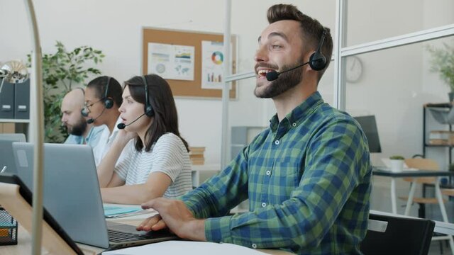 Tired guy is working in call center talking and typing then putting head on table relaxing feeling exhausted while colleagues are concentrated on work in background